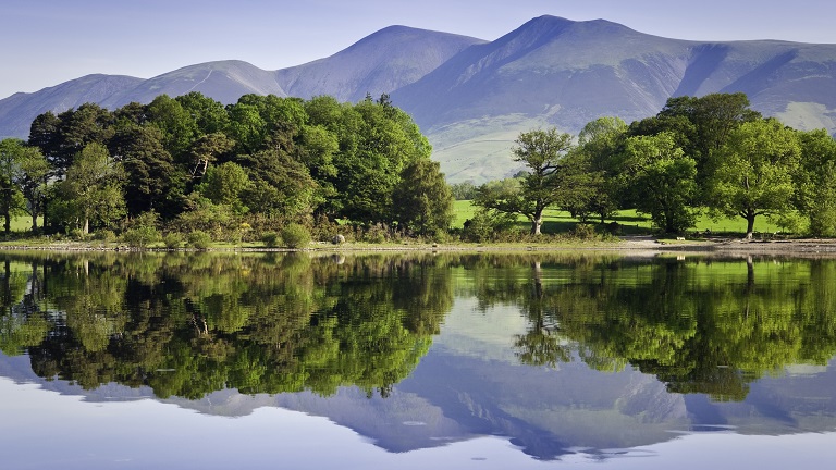 Trees and mountains reflected in the mirror-like waters of Derwentwater, a popular lake in the Lake District National Park